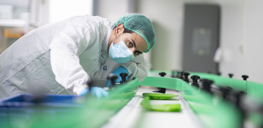 A quality control technician in a lab coat and mask inspects products on a manufacturing line.
